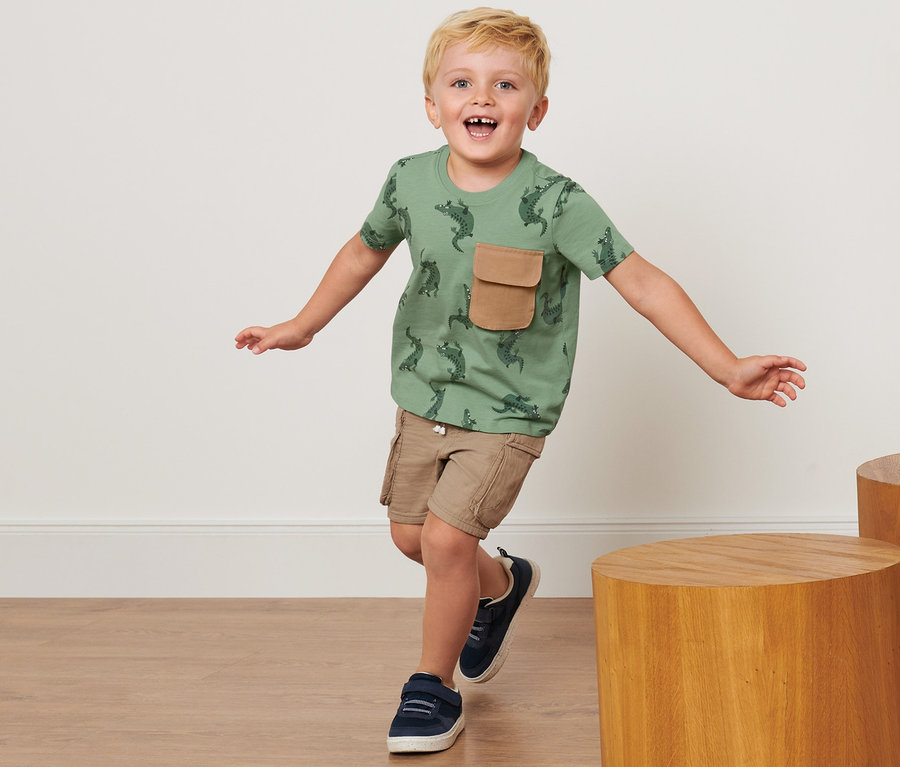 Ein Junge in einem Kinder-T-Shirt mit Krokodilmuster und einer braunen Tasche rennt drinnen.