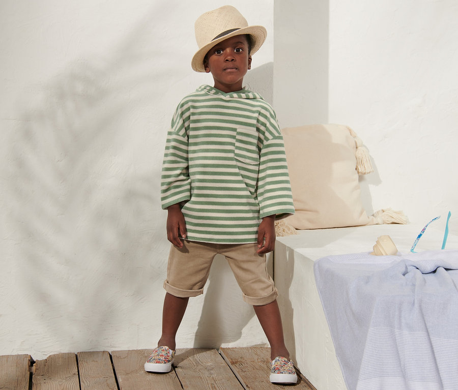 Junge in Kinder-Frottier-Shirt, beigen Shorts und Hut steht an einer weißen Wand.