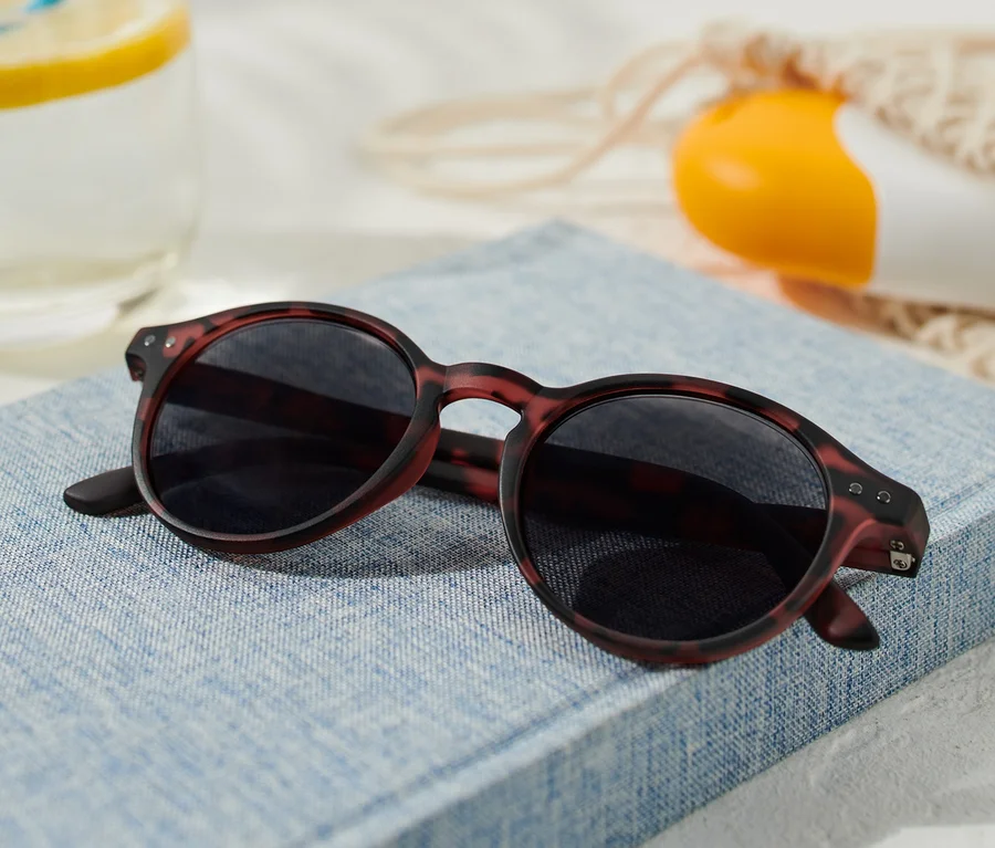 Eine getönte Lesebrille liegt auf einem blauen Buch, neben einem Glas mit Zitrone und einer Sonnencreme.