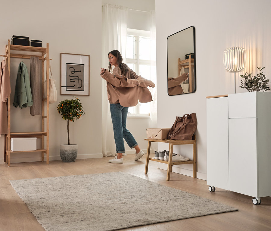 Eine Frau zieht im Flur eine Jacke an. Im Hintergrund befinden sich ein Highboard auf Rollen, eine Standgarderobe mit Ablageflächen, eine Tischleuchte aus Metall und ein Hocker mit breiten Ablageflächen.