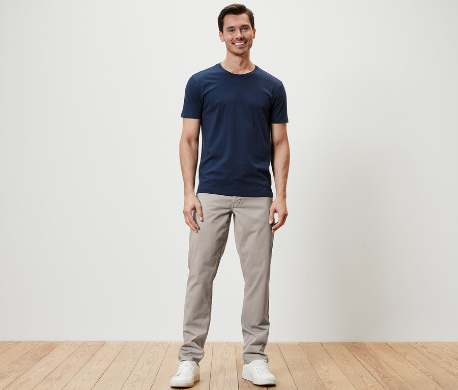 Mann steht und trägt eine 5-Pocket-Hose, ein T-Shirt und Turnschuhe.