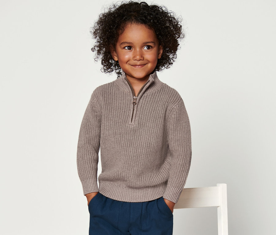 Lächelnder Junge posiert in braunem Kinder-Strickpullover, Troyer, blauen Hosen und grünen Socken.