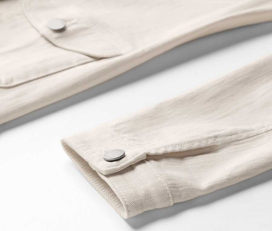 Detailaufnahme einer hellbeigen Jeansjacke mit Cargo-Taschen, die eine Brusttasche und eine Ärmelbündchen zeigt.