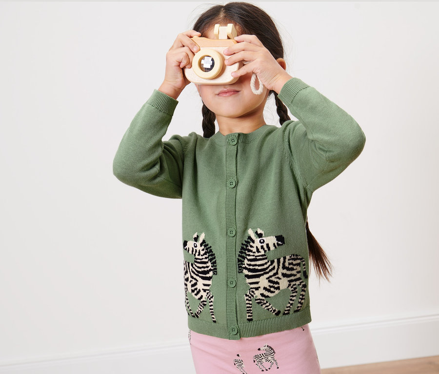 Mädchen mit Zöpfen hält eine Holzkamera vor ihr Gesicht und trägt eine grüne Kinder-Jacquard-Strickjacke mit Zebra-Motiv.