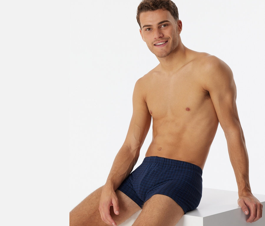 Mann sitzt auf einem weißen Würfel und trägt karierte SCHIESSER Shorts.