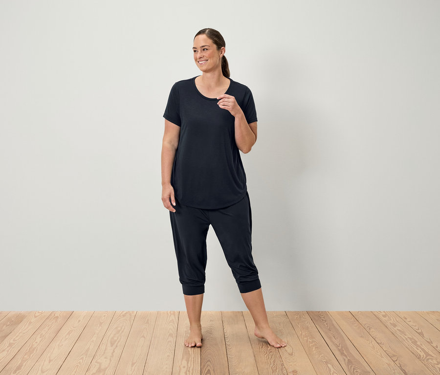 Eine Frau in einem schwarzen T-Shirt und Dreiviertelhosen steht auf einem Holzboden vor einer weißen Wand.