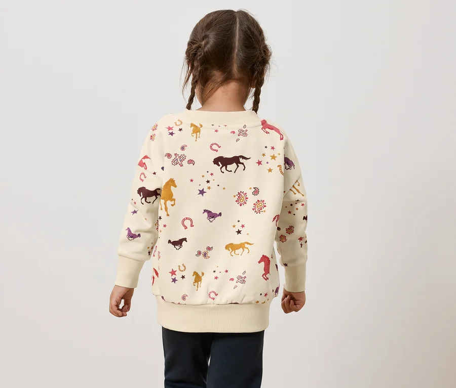 Mädchen mit Zöpfen von hinten trägt ein cremefarbenes Sweatshirt mit Pferde-, Hufeisen- und Sternenmuster.