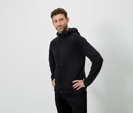 Ein Mann posiert vor einem weißen Hintergrund. Er trägt einen schwarzen Kapuzenpullover und eine schwarze Hose.