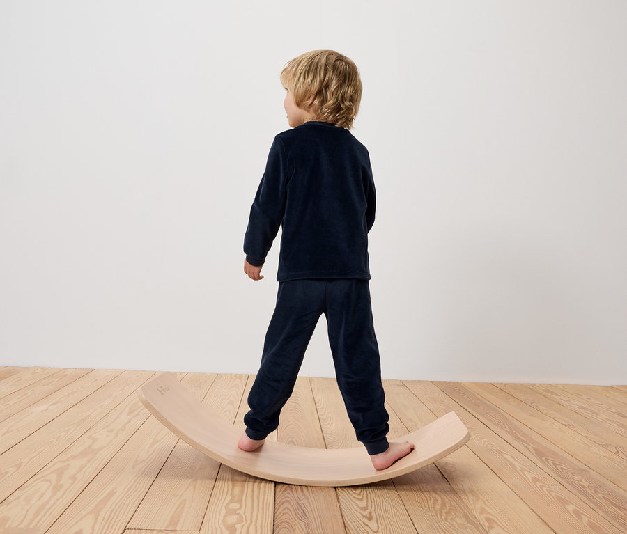 Junge in blauem Schlafanzug steht auf einem hölzernen Balance Board.