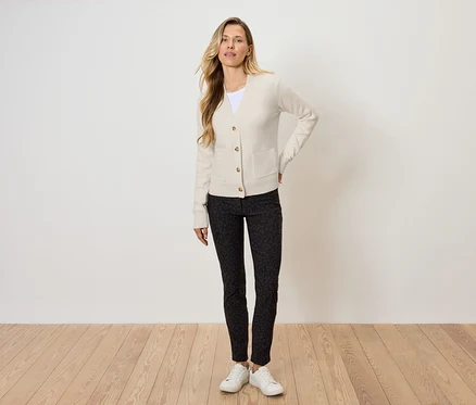Blonde Frau posiert in cremefarbenem Cardigan, gemusterter, schwarzer Hose und weißen Turnschuhen.