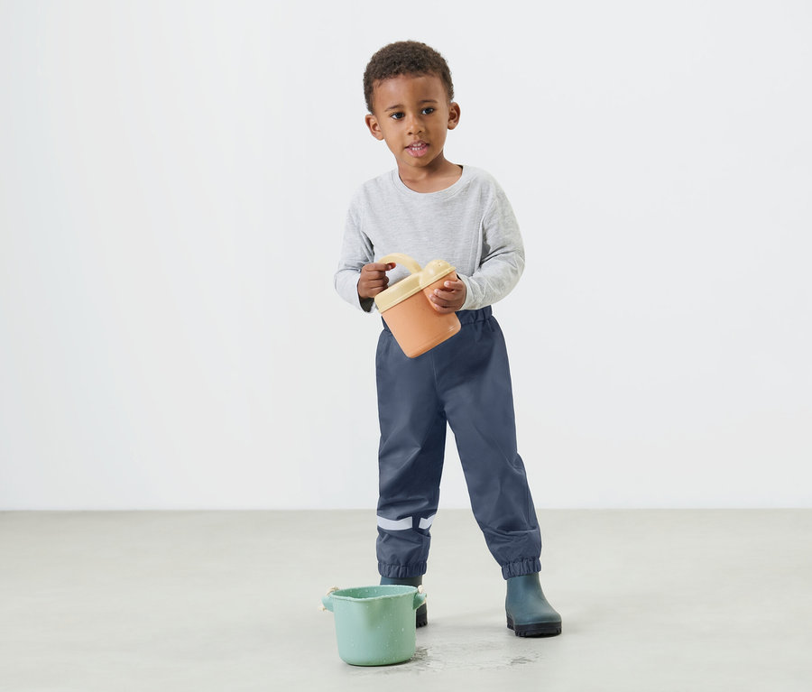 Ein kleiner Junge hält einen orangefarbenen Eimer in seinen Händen und trägt dabei blaue Regenhosen und Gummistiefel.
