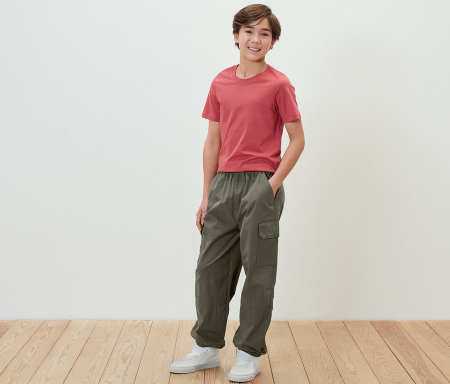 Ein Junge in einem roten T-Shirt und einer Kinder-Cargohose steht vor einer weißen Wand.