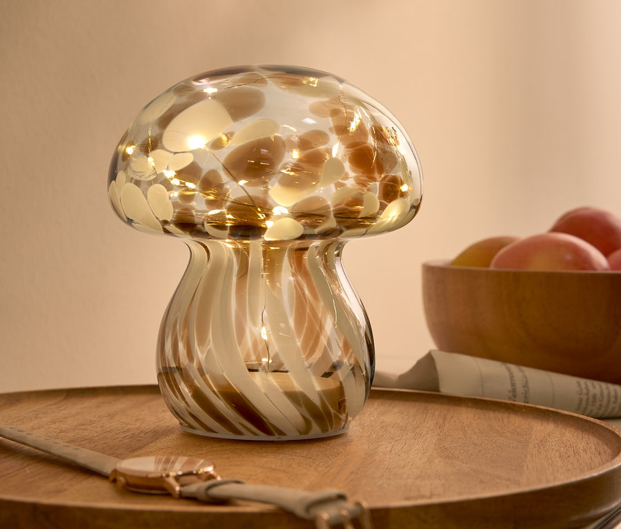 Dekorative Glaslampe in Form eines Pilzes mit braunen Flecken und Lichtern, auf einem Holztablett neben einer Uhr und einer Schale mit Früchten.
