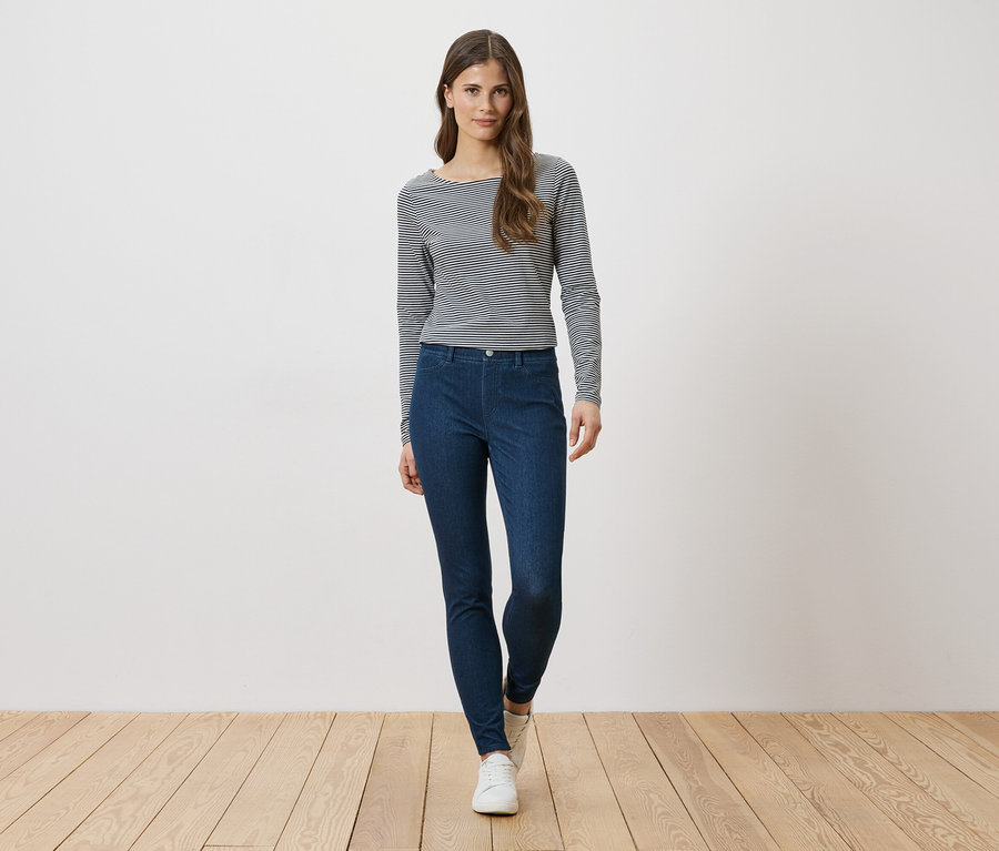 Frau mit langärmligem, gestreiftem Oberteil und blauen Jeans, die vor einer weißen Wand steht.