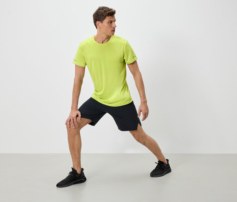 Mann in Sportbekleidung dehnt sich. Er trägt ein neon-grünes T-Shirt, schwarze Shorts und schwarze Turnschuhe.