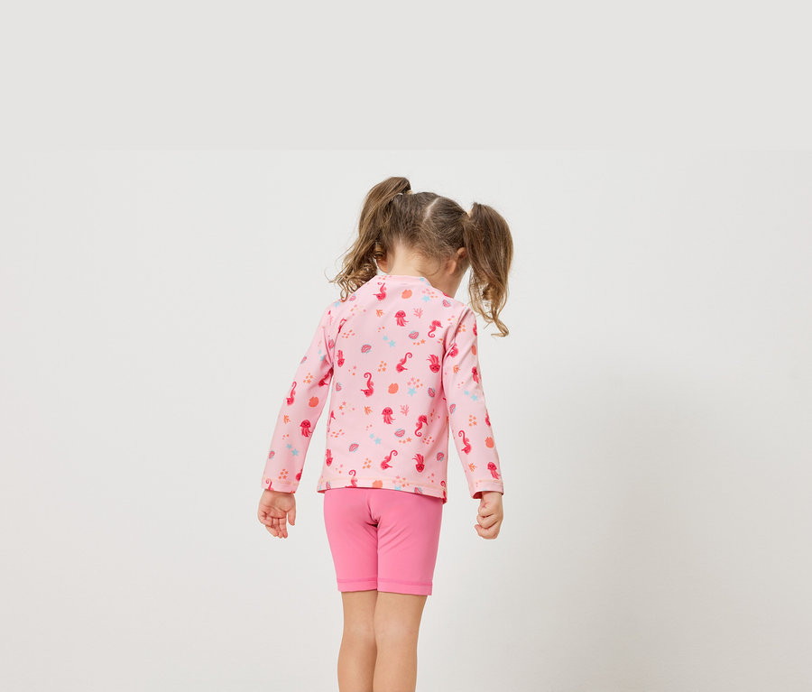 Ein kleines Mädchen mit Zöpfen trägt ein rosa Langarmshirt mit Meeresmotiven und rosafarbene Shorts.