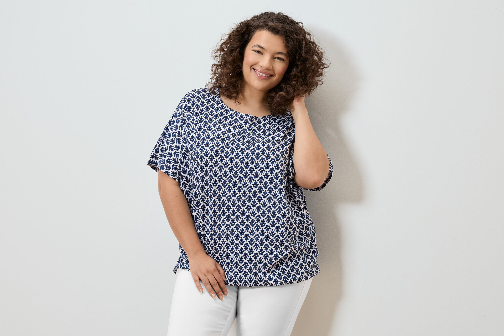 Frau mit lockigem Haar, die eine blau-weiße Bluse und weiße Hosen trägt und lächelt.