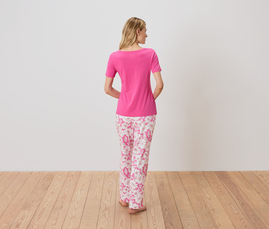 Rückansicht einer Frau mit blonden Haaren, die ein pinkes T-Shirt und eine rosafarbene Pyjamahose mit Blumenmuster auf einem Holzboden trägt.