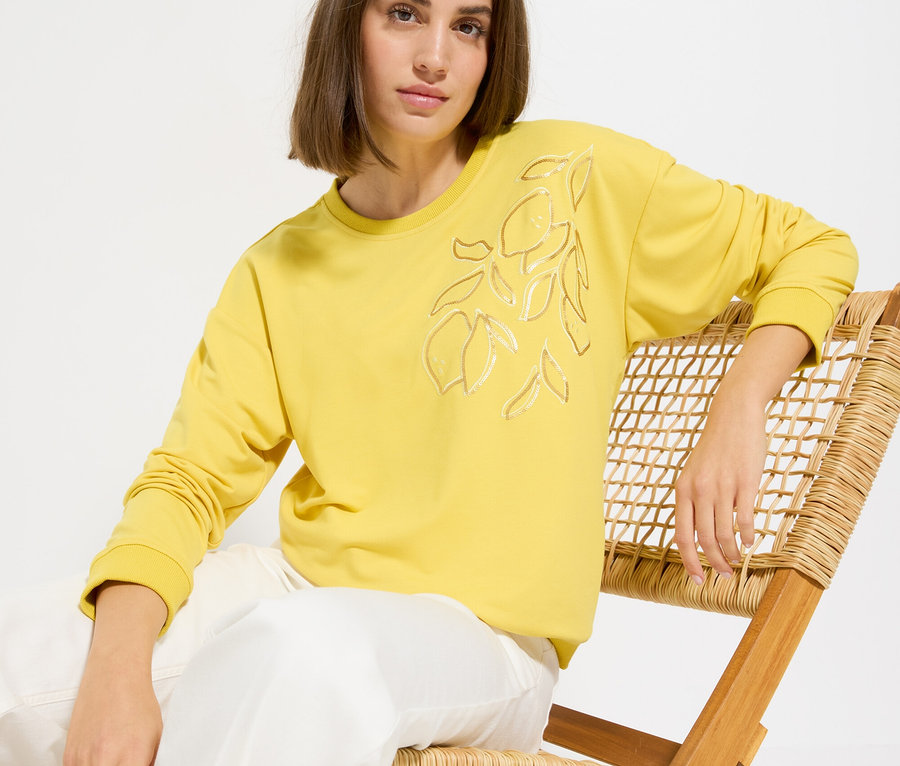 Frau sitzt auf einem Stuhl und trägt einen gelben Pullover mit Zitronenmuster und eine weiße Hose.