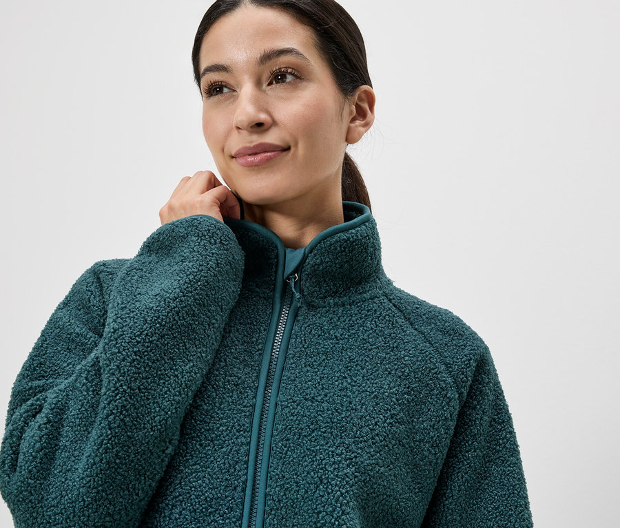 Frau mit Zopf trägt eine grüne Fleecejacke mit Reißverschluss und hohem Kragen.