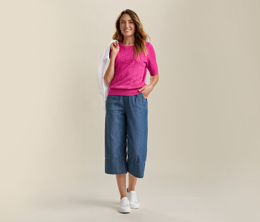 Frau mit Jeansjacke mit Lochstickerei und pinkem Feinstrickpullover mit Zopfmuster. Sie trägt blaue Culottes und weiße Sneaker.