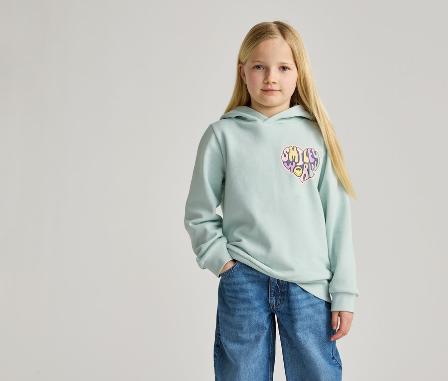 Mädchen mit blonden Haaren trägt hellblauen Kapuzenpullover mit Herz-Smiley-Aufdruck und Jeans.