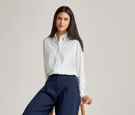 Frau mit dunklen Haaren, trägt eine weiße Bluse mit schwarzen Punkten und eine blaue Hose, während sie auf einem Hocker sitzt.