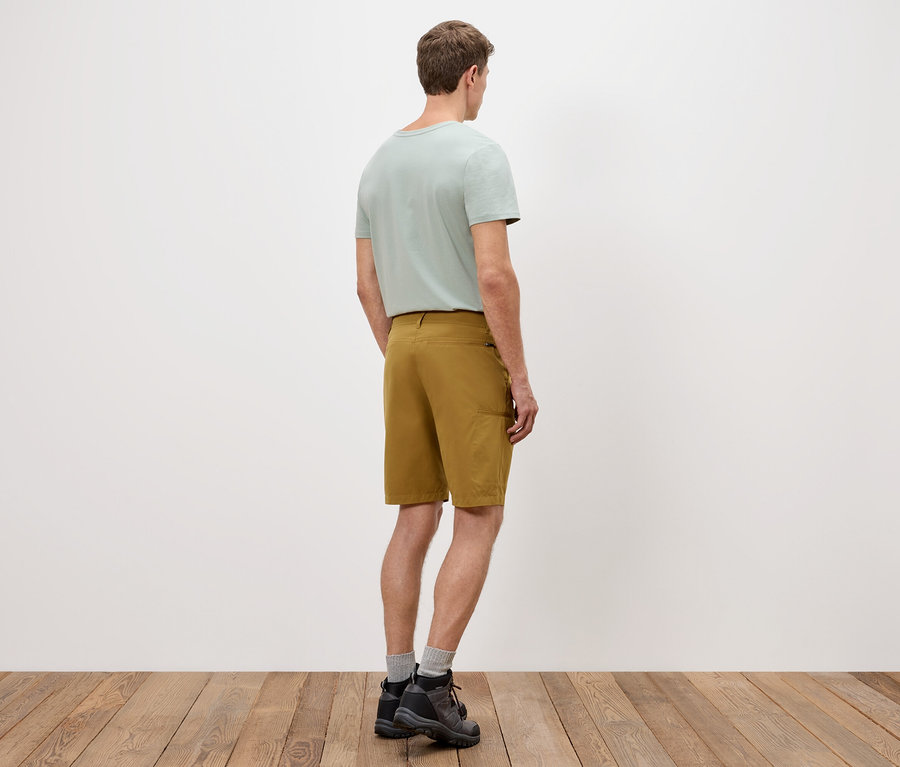 Ein Mann steht mit dem Rücken zur Kamera, trägt ein hellgrünes T-Shirt, goldene Shorts und dunkle Schuhe auf einem Holzboden.
