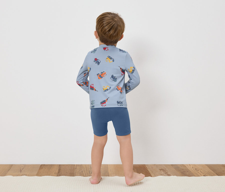 Ein Kind steht mit dem Rücken zur Kamera, es trägt ein blaues Langarmshirt mit bunten Baufahrzeugen und blaue Shorts.