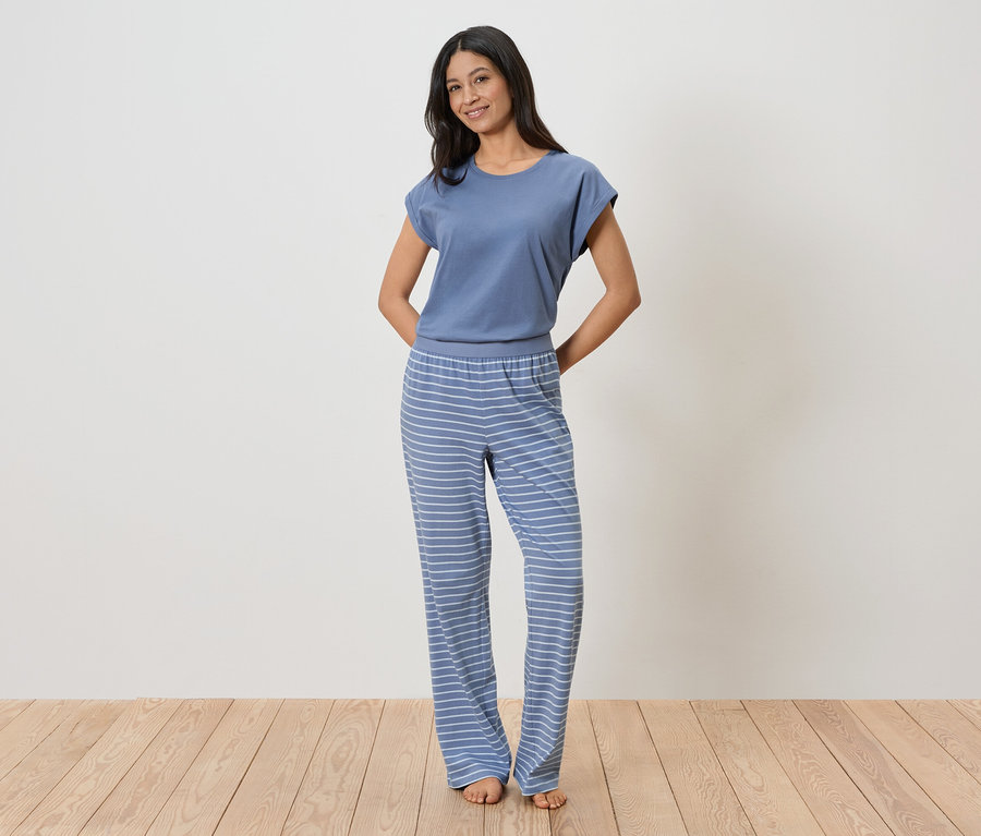 Frau mit blauem T-Shirt und gestreifter Hose steht vor einer weißen Wand.