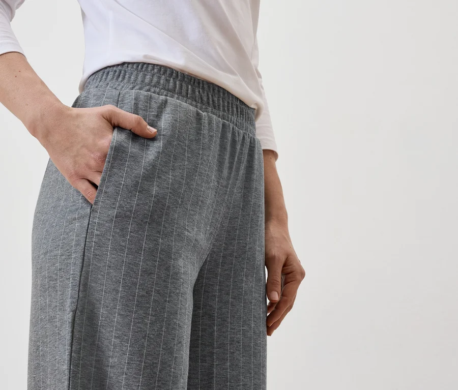 Eine Person mit einer Hand in der Tasche einer grauen, gestreiften Hose, kombiniert mit einem weißen Oberteil.