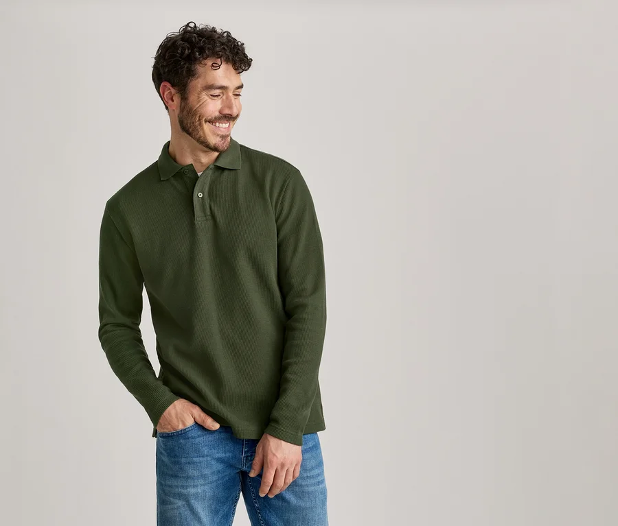 Lächelnder Mann mit lockigem Haar, der ein olivgrünes Langarmpolo und Jeans trägt, mit einer Hand in der Hosentasche.