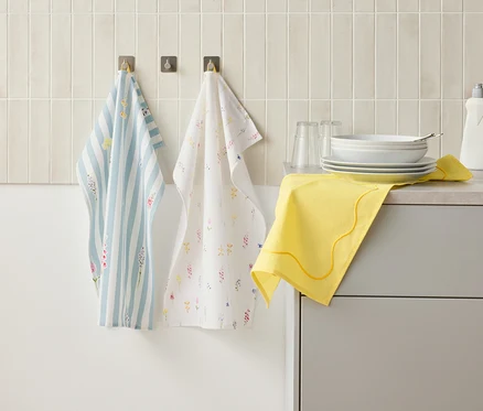 Zwei Geschirrtücher hängen an Haken an einer Wand über einer Küchentheke, auf der sich Geschirr, Gläser, eine Spülmittelflasche und ein gelbes Tuch befinden.