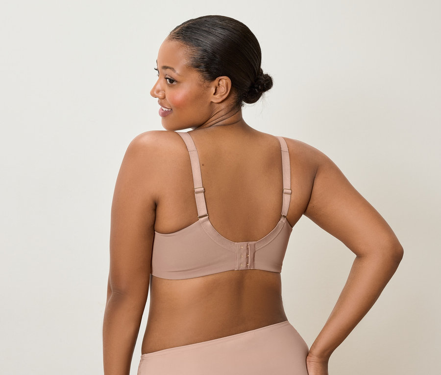 Rückansicht einer Frau mit dunkler Haut und Dutt, die einen nudefarbenen BH und passende Unterwäsche trägt und über die Schulter nach links blickt.