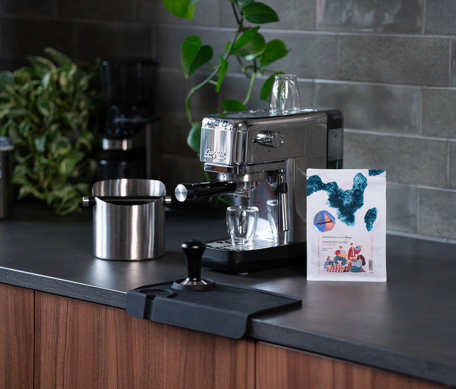 Eine silberne Espressomaschine steht auf einer Küchentheke neben einer Packung Roasted Community Blend Kaffee.