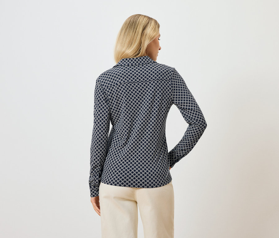 Rückansicht einer blonden Frau, die eine langärmlige, blau gemusterte Bluse und eine helle Hose trägt.