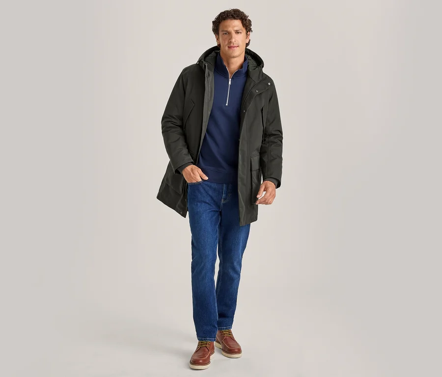 Mann steht, trägt einen dunkelgrünen Kapuzenparka, einen marineblauen Quarter-Zip-Pullover, blaue Jeans und braune Bootsschuhe, Hände in den Taschen.