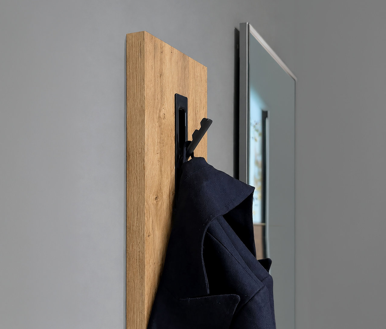 Holzpaneel mit schwarzem Garderobenhaken an grauer Wand, daran hängt ein dunkelblauer Mantel. Rechts ist ein Spiegel teilweise sichtbar.