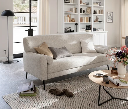 Helles Wohnzimmer mit einem beigen Sofa, das mit beigen, grauen und weißen Kissen dekoriert ist. Vor dem Sofa steht ein runder Holztisch mit einer Tasse, einer Zeitschrift und einer Vase mit bunten Blumen. Auf dem gemusterten Teppich liegen Hausschuhe. Im Hintergrund sind ein Bücherregal und ein großes Fenster zu sehen.