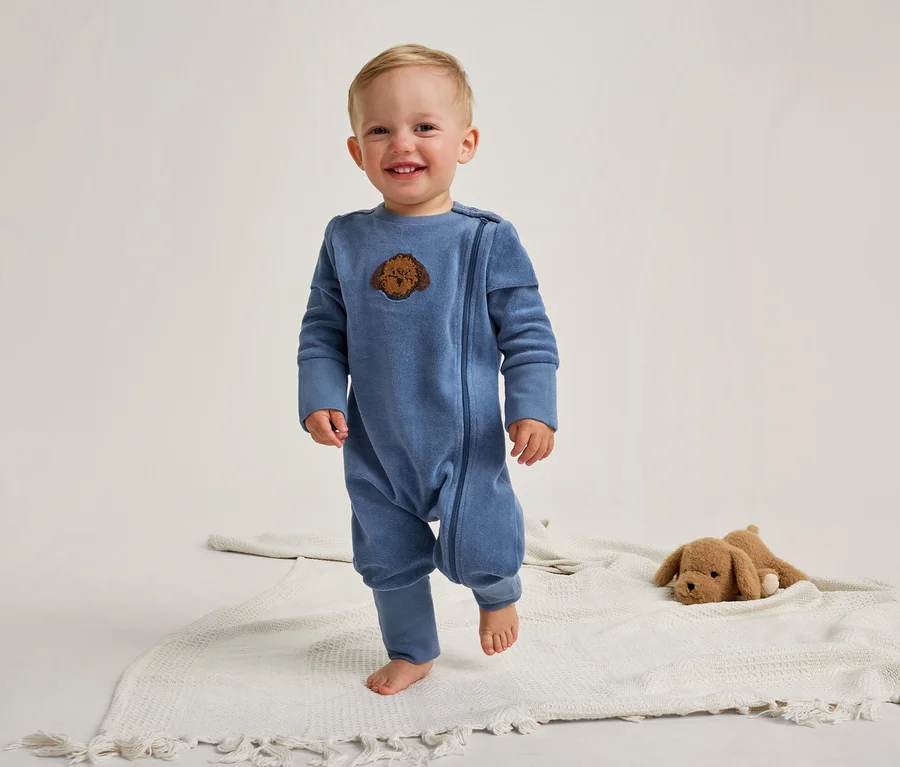 Ein lächelndes Baby steht barfuß auf einer weißen Decke. Es trägt einen blauen Strampler mit einem braunen Hundekopf-Aufnäher. Rechts liegt ein braunes Plüschtier.
