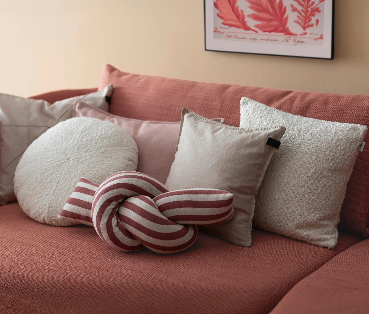 Ein terrakottafarbenes Sofa mit verschiedenen Kissen, darunter ein rot-weiß gestreiftes Knotenkissen »JOLLY«, ein rundes cremefarbenes Kissen und mehrere rechteckige beige und cremefarbene Kissen.
