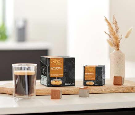 Ein Glas Kaffee, eine 27er- und eine 8er-Packung Qbo Caffè BUNA ENTETA Kapseln sowie zwei einzelne Qbo Kapseln stehen auf einem Holzbrett. Eine Schale mit Keksen und eine Vase mit Trockenblumen sind im Hintergrund.