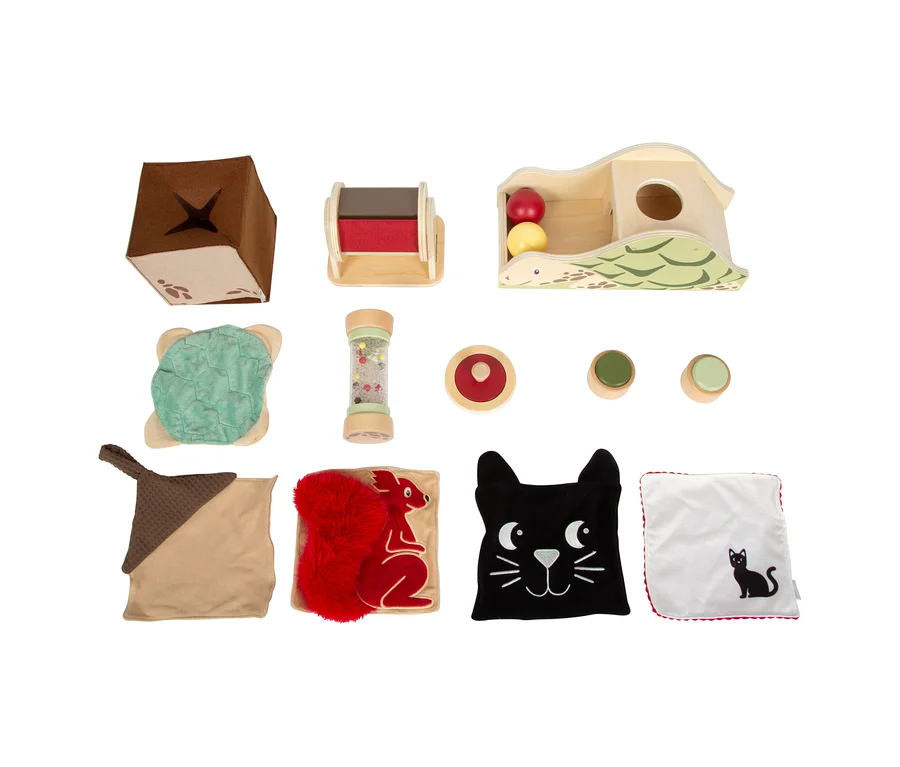 Eine Anordnung von zehn Spielzeugen aus der small foot Baby-Box für 7–9 Monate, darunter ein brauner Stoffwürfel, ein Holzroller, ein Holzspielzeug in Schildkrötenform mit zwei Bällen, ein grüner Stoffschildkrötenpanzer, eine Rassel, drei Holzscheiben und vier Stofftücher mit Tiermotiven.