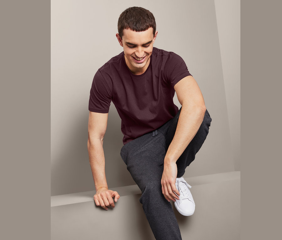 Ein Mann sitzt, trägt ein weinrotes T-Shirt und eine graue Trainingshose.