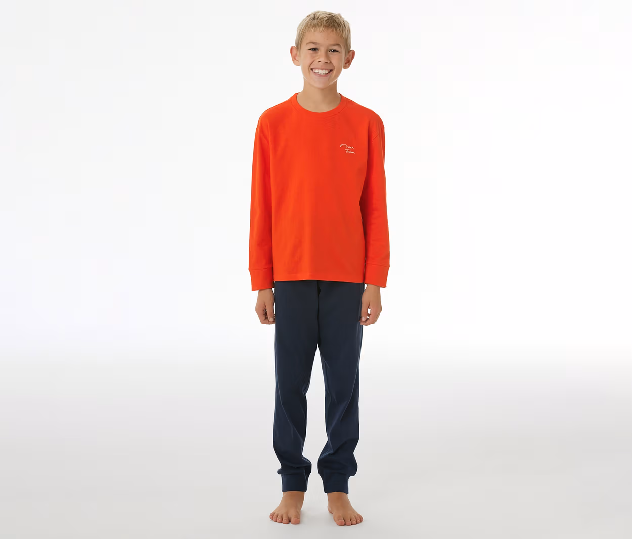 Lächelnder Junge in einem orangefarbenen SCHIESSER Kinder-Pyjama mit langen Ärmeln und dunkelblauer Hose mit Bündchen.