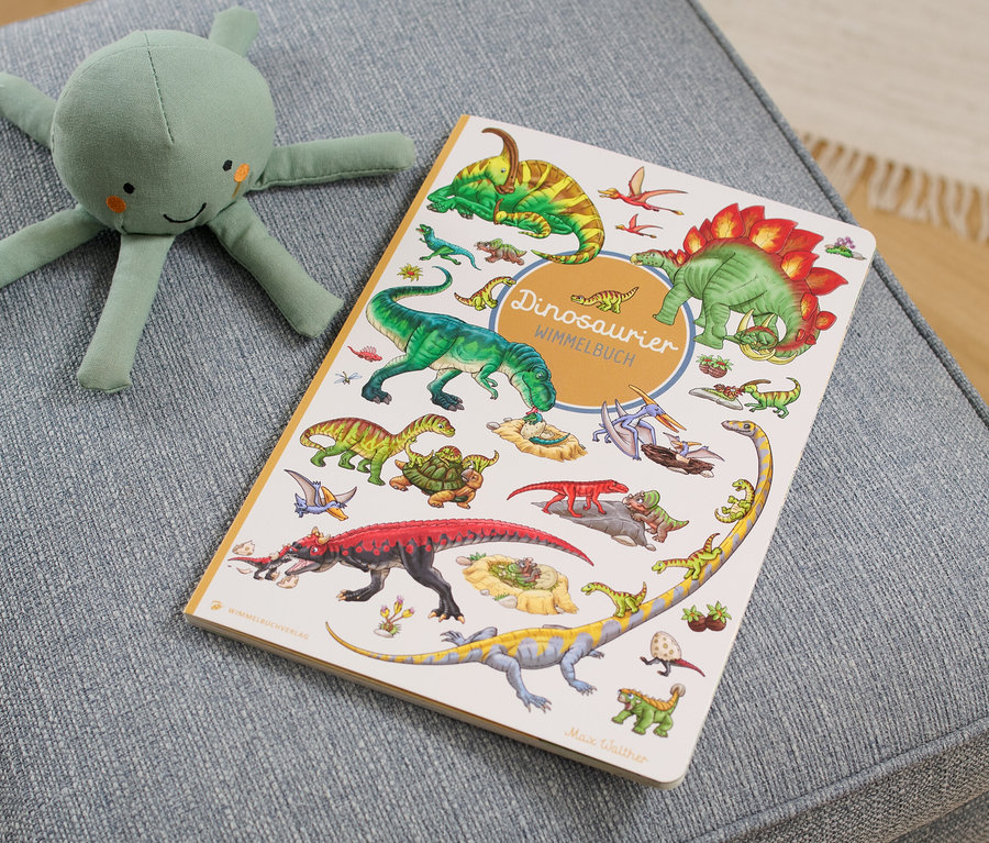 Auf einem grauen Hocker liegen ein »Dinosaurier Wimmelbuch« und ein grünes Tintenfisch Stofftier.