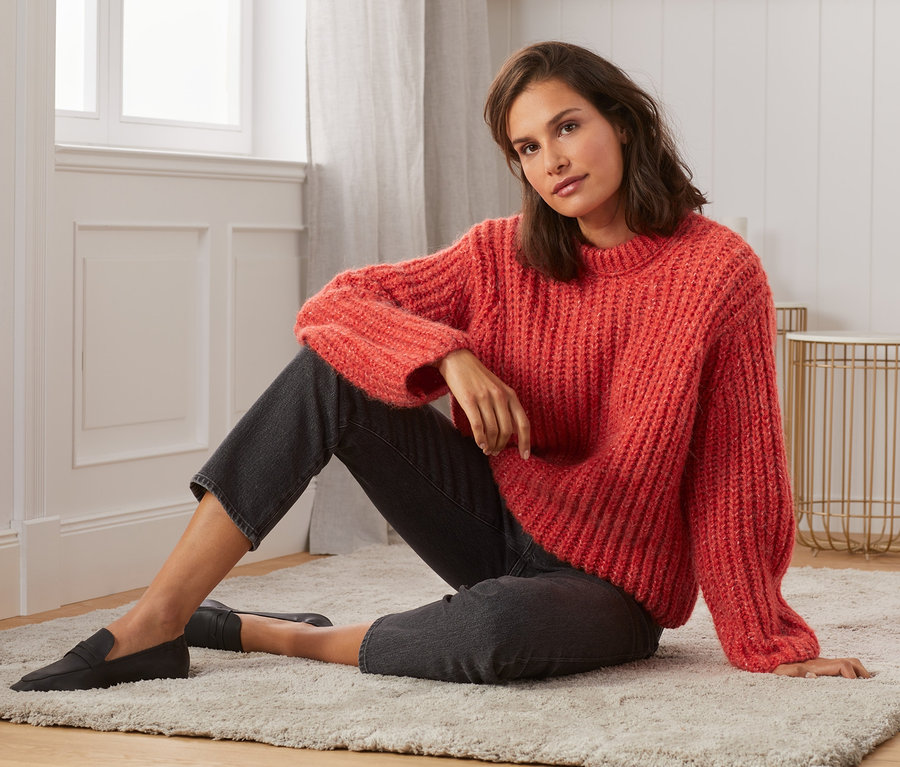 Eine Frau sitzt auf einem Teppich und trägt einen Grobstrickpullover.