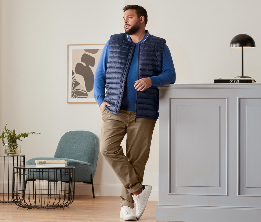 Mann lehnt an Kommode, trägt blaue Steppweste, royalblauen Leinen-Pullover, camelfarbene Chino und Sneaker.