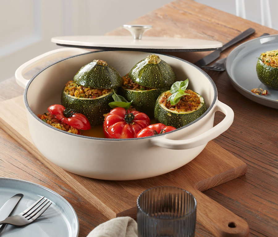 Auf einem Holztisch steht eine cremefarbene Gusseisen-Bräter-Pfanne mit gefüllten Zucchini und Tomaten.