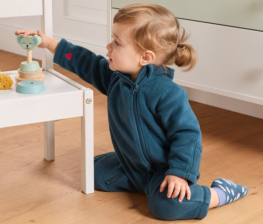 Kleinkind spielt mit Holzspielzeug auf einem weißen Stuhl, es trägt einen Fleece-Einteiler aus recyceltem Material.
